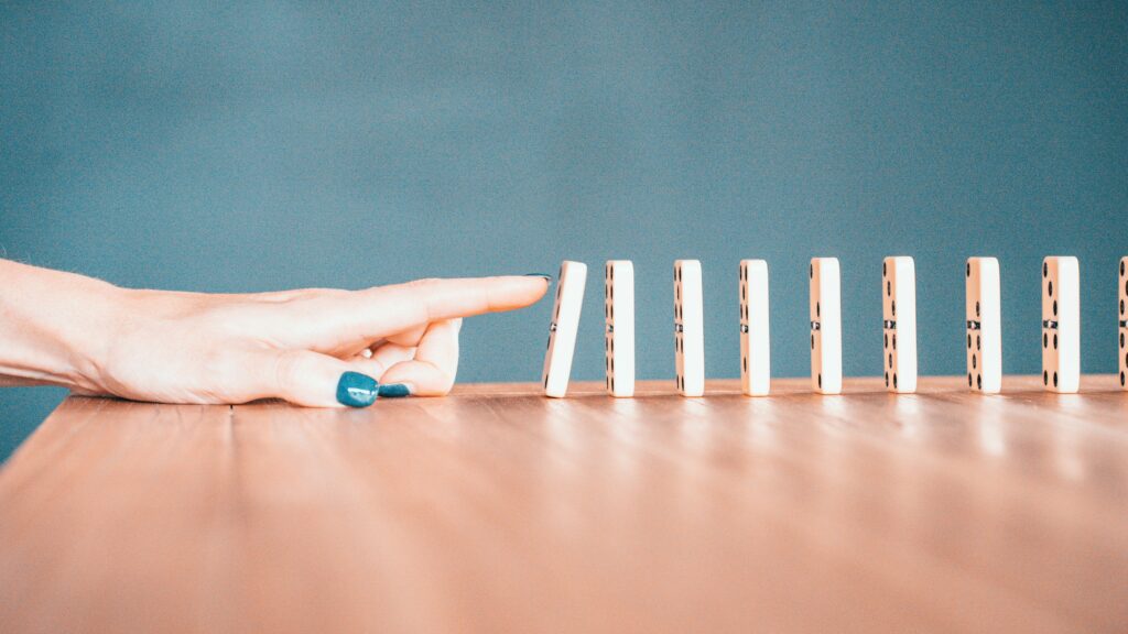 Weiße Dominosteine auf einer Tischplatte hintereinander aufgestellt, eine Hand mit blau lackierten Fingernägeln tippt den ersten Stein an, der sich bereits zum Umfallen neigt.